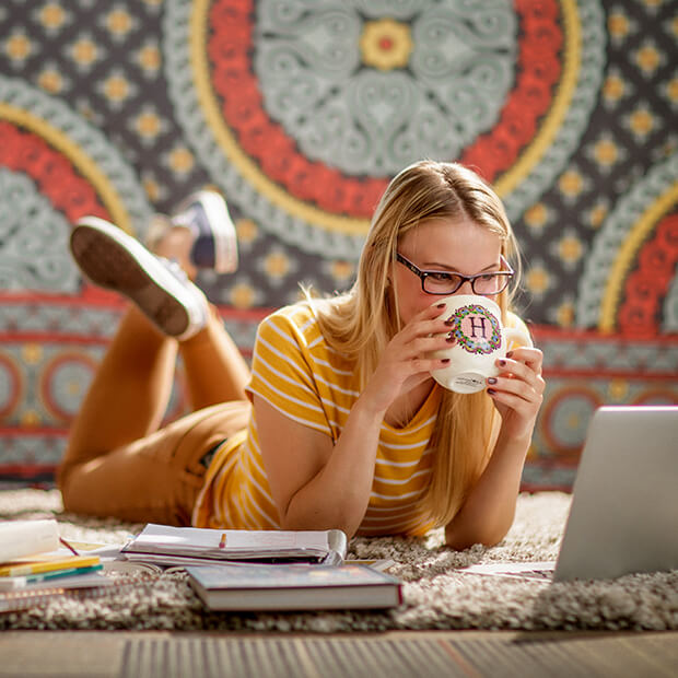 Women laying on the floor, looking at her laptop, whilst drinking coffee