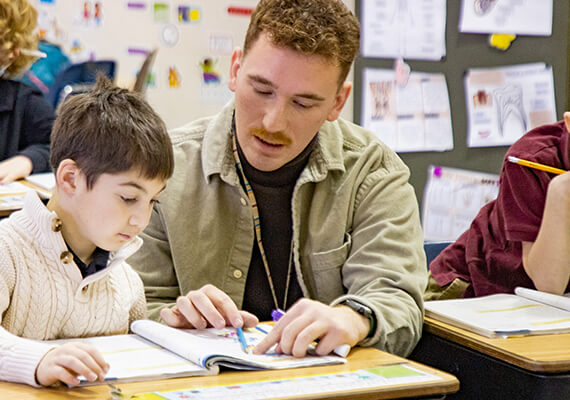 A schoolteacher kneels next to a grade school student and explains a math problem